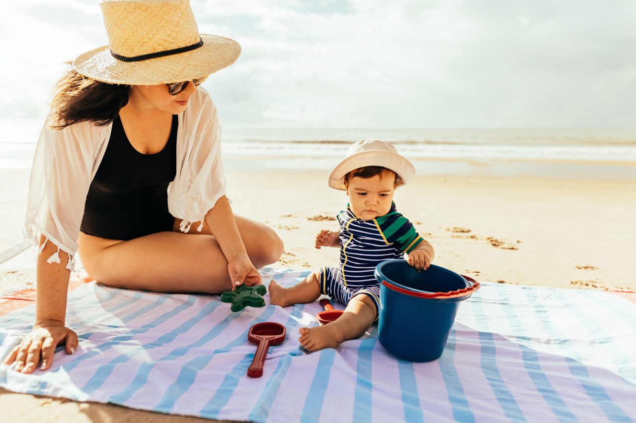 Kleinkind und Mutter spielen im Urlaub auf Decke im Sand
