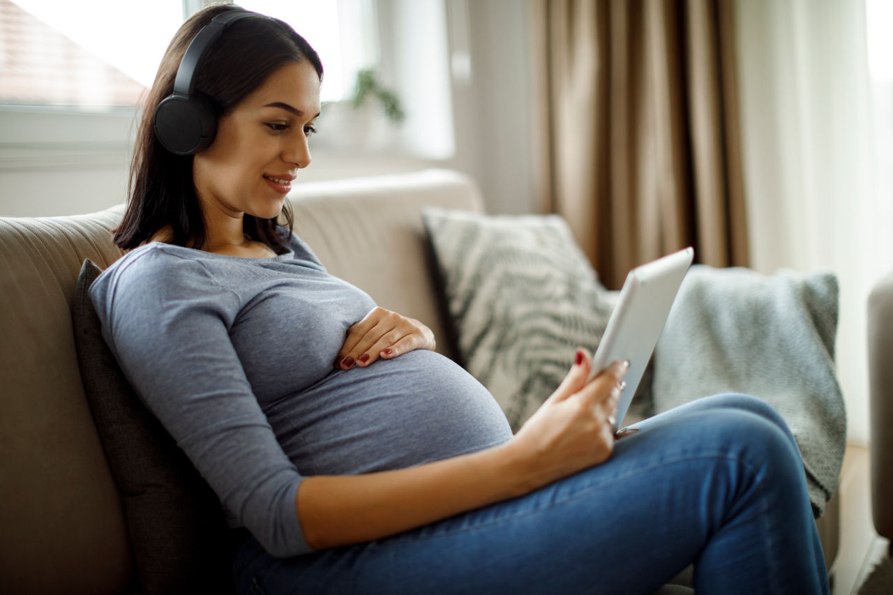 Smiling pregnant woman with bluetooth headphones using digital tablet at home