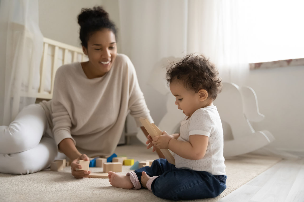 Happy young biracial mother sit on floor play with building blocks bricks with little infant toddler, smiling african American mom engaged in learning activity with small baby child at home